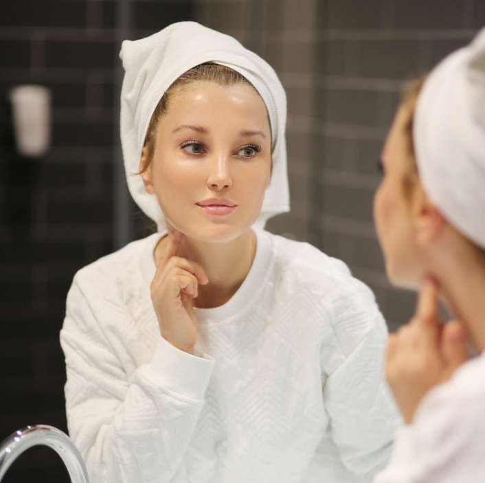 A woman in a white bathrobe applying Golden Peptide Lifting Cream while looking in the mirror.