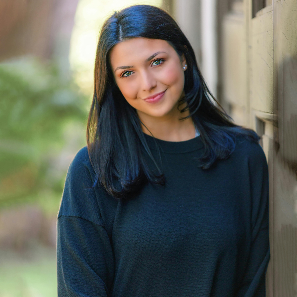 Close-up of a woman with beautiful skin, wearing a black sweater, smiling outdoors.