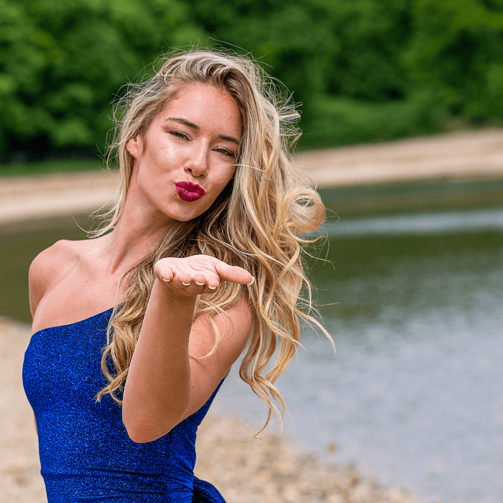 Woman with flowing blonde hair blowing a kiss, wearing a blue dress near a serene lakeside.