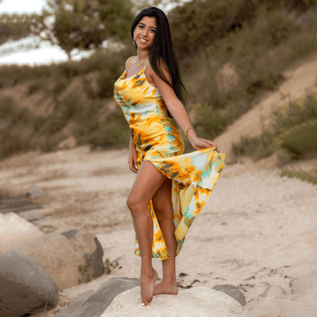 A woman wearing a colorful yellow and turquoise dress, posing on a beach with a relaxed smile.