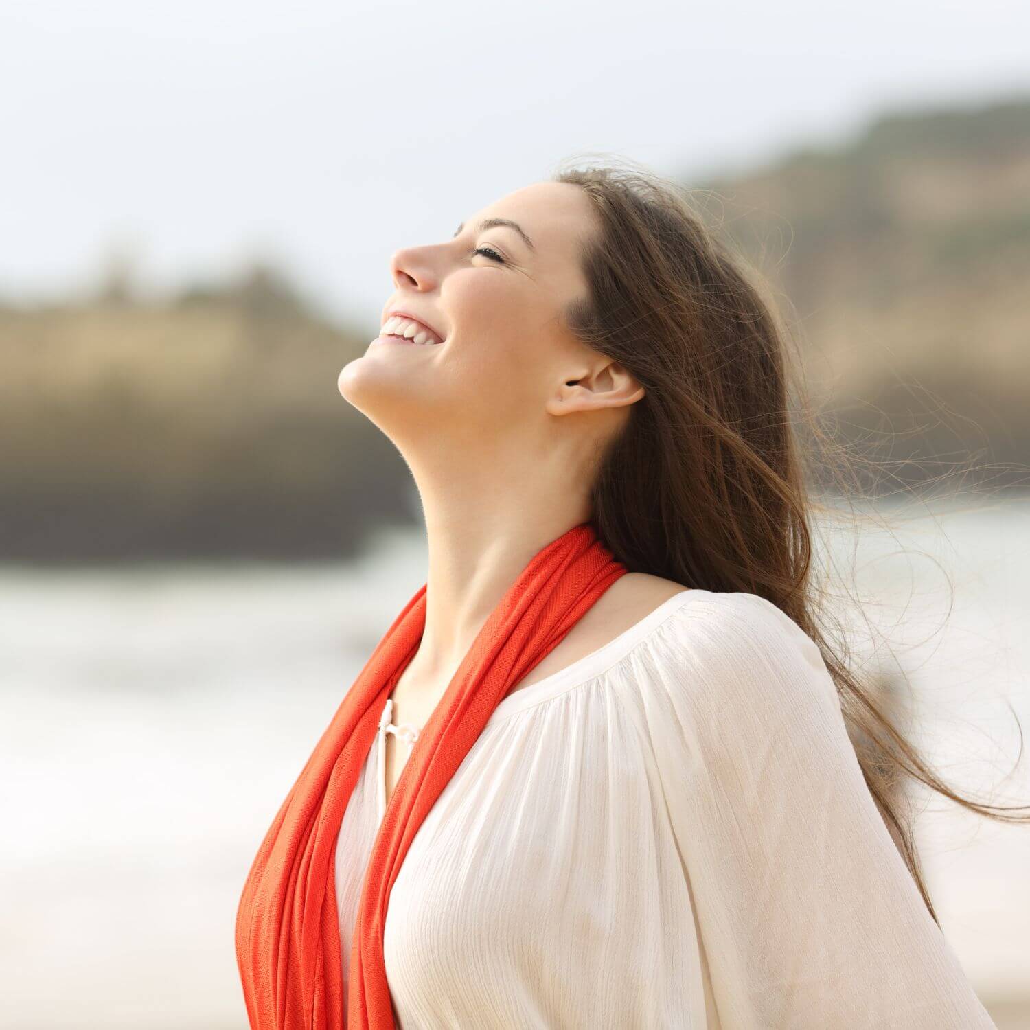 Smiling woman with long hair wearing a white blouse and orange scarf by the beach, exuding happiness and tranquility.
